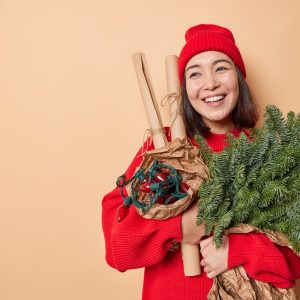 Happy dreamy young Asian woman thinks about New Year celebration holds green spruce branches rolled paper and retro garland prepares for holidays isolated over beige background with blank space